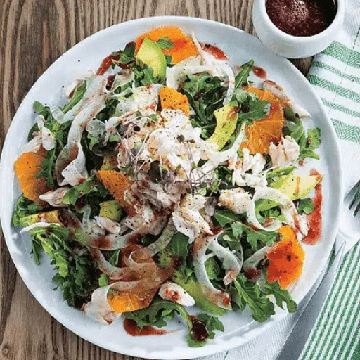 A vibrant salad with arugula, shredded chicken, orange slices, avocado, and microgreens, drizzled with dark vinaigrette, served on a wooden table.
