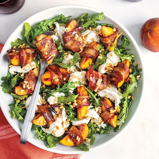 A fresh salad with arugula, burrata cheese, grilled peach slices wrapped in bacon, topped with pine nuts. The setting is bright and inviting.
